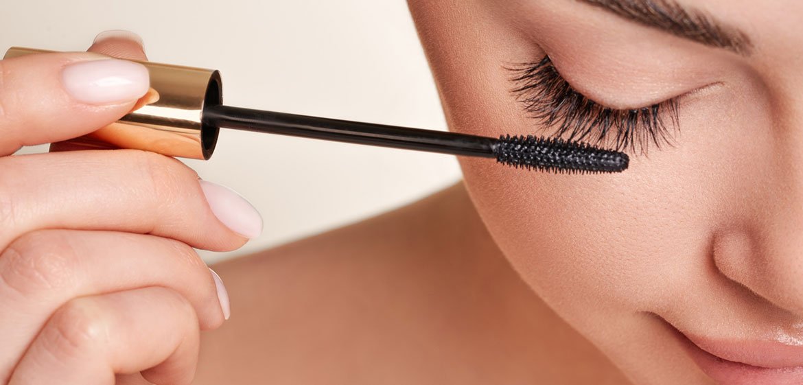 Eine junge Frau hält einen Mascara an ihre Wimpern und schaut mit geschlossenen Augen nach unten.